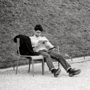 A man sits alone on a chair outdoors, reading a book. He is wearing glasses, jeans, and a jumper, with a jacket draped over the chair beside him. The background is a grassy slope.