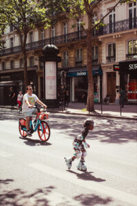 A man rides a hire bike while a child wearing roller pads and a helmet skates ahead of him on a sunny city street lined with shops and trees.