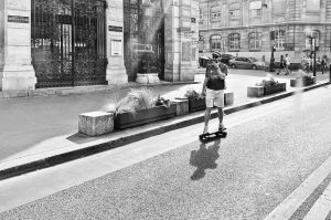 A person rides a skateboard down a city street on a sunny day, wearing shorts, a vest, helmet, and sunglasses. Historic buildings and parked cars are visible in the background. The scene is in black and white.