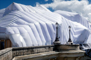 A large building is covered in a white, inflated plastic sheet, creating a billowing effect. Ornate street lamps stand in front, and part of a stone balustrade is visible under a partly cloudy sky.