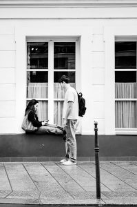 Jacob & Rosaline share a quiet moment—a young woman sits on a windowsill looking at her mobile as Jacob, wearing a rucksack, stands nearby. Both are outside a building with large windows and flowing curtains.