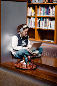 Bernadette Derrien, wearing headphones, sits on wooden steps in a library, focused on her laptop. Nearby are a brown bag and green water bottle, with bookcases in the background.