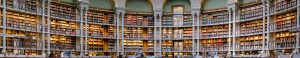 A panoramic view of a grand, circular library with multiple levels of bookshelves, ornate arches, and rows of tables and chairs for reading and studying. Warm lighting illuminates the library’s vast collection of books.