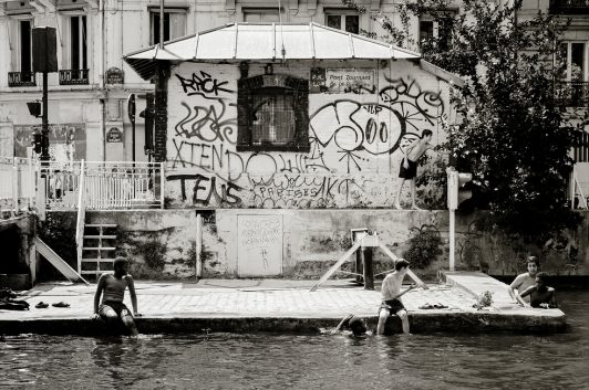 Three people sit by the edge of a canal, dipping their feet in the water. Behind them is a small, graffiti-covered building and some trees. The scene has a relaxed, urban atmosphere. Image is black and white.