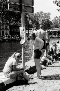 A group of people in swimwear gather beside a canal on a sunny day. One woman sits on a bag looking at her mobile, while others stand or sit near the water. Trees and a poster on a pole are visible in the background.