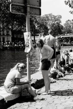 A group of people in swimwear gather beside a canal on a sunny day. One woman sits on a bag looking at her mobile, while others stand or sit near the water. Trees and a poster on a pole are visible in the background.
