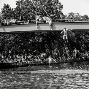 Black and white photo of two people jumping off a bridge into a river, with crowds watching from the bridge and riverbank. Trees fill the background.