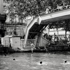 A black and white photo of people gathered around and swimming in a canal, whilst one person jumps into the water from a footbridge overhead. Trees and buildings are visible in the background.