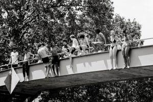 A group of young people in swimming costumes sit and stand on the edge of a bridge, preparing to jump into the water below. Trees fill the background on a sunny day. The image is in black and white.