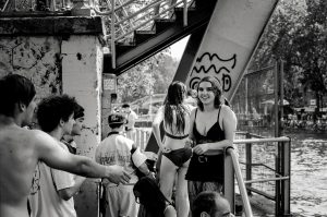 A group of people in swimsuits gather by a canal under a staircase. One woman in front, wearing a black swimming costume, smiles at the camera while others chat and enjoy the water. The scene is lively and relaxed. Black and white photo.
