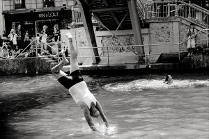 A man in a vest and shorts dives headfirst into a canal, while another swimmer is in the water nearby; people watch from a bridge and riverside in a city setting. The photo is in black and white.