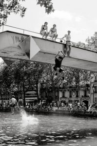 A black-and-white photo shows several people on a bridge over a canal; one person is jumping off the bridge into the water below whilst others watch. People are gathered along the bank, and trees line the background.