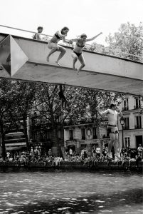 Three people in swimming costumes jump off a bridge into a canal, whilst crowds of people watch from the water’s edge on a sunny day. The scene is in black and white, with trees and buildings in the background.