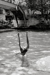 A person is captured mid-air upside down with legs up, diving or falling into a canal whilst people watch from a footbridge and the bank in a city setting. The image is in black and white.