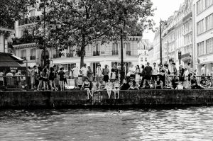 A large group of people sit and stand along the edge of a city canal, some dangling their feet over the water, whilst one person swims. The scene is lively, set against buildings and trees, in black and white.