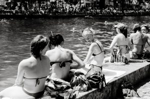 A group of women in swimsuits sit on a sunny riverside, talking and relaxing. Bags and towels are beside them, whilst people gather on the opposite riverbank in the background. The image is in black and white.