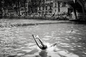 A person dives upside down into a canal, splashing water, whilst another swims nearby. A crowd sits along the edge, watching, with buildings and trees lining the background. The image is in black and white.