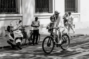 A man wearing a hat rides a bicycle with a young boy sitting in front, both wearing helmets, on a city street. In the background, two people walk by and another stands near parked motorbikes and bikes. The image is in black and white.
