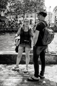 A woman and a man stand talking beside a canal in a city. The woman wears shorts, glasses, and a camera round her neck; the man wears a cap and rucksack. People sit in the background by the water. The photo is black and white.
