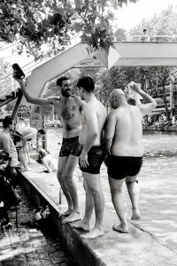Three men in swimwear stand on a riverbank under a bridge, talking and laughing. Other people are swimming and relaxing by the water in the background. The image is in black and white.