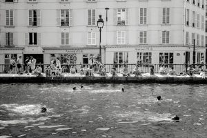 A black and white photo shows people sitting along a canal edge, while several swimmers are in the water. Behind them is a building with many windows and French signage. The scene appears lively and relaxed.