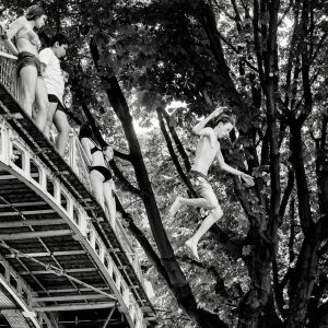 Four young people stand on a metal bridge whilst one person leaps off into the air, surrounded by leafy trees, in a black and white photo capturing an adventurous summer moment.