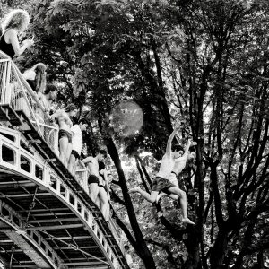 A group of people stand on a bridge, some watching and some preparing to jump. One person is captured mid-air leaping off the bridge, surrounded by leafy trees on a sunny day. The image is in black and white.