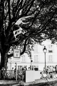 A person jumps from a tree over a canal, arms outstretched, while a crowd watches from the canal’s edge below. The scene is in black and white with large trees and buildings in the background.
