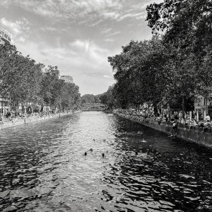 Black and white photo of a wide canal lined with trees and people sitting along both banks. Some people are swimming in the water, and the sky is partly cloudy. A bridge is visible in the distance.