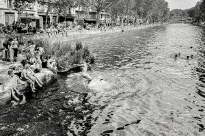 Black and white photo of people relaxing and swimming along the banks of a canal in a city, with some individuals jumping into the water and others sitting or sunbathing near the edge.