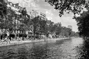 Black and white photo of a canal lined with trees and buildings, with people sitting, walking, and relaxing along the water’s edge. Swimmers are visible in the canal, and “HÔTEL DU NORD” is painted on one building.