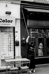 A person stands outside a restaurant, using a long pole to reach something above a shop entrance. Empty tables and chairs are in the foreground; a menu is visible on the wall. The image is in black and white.