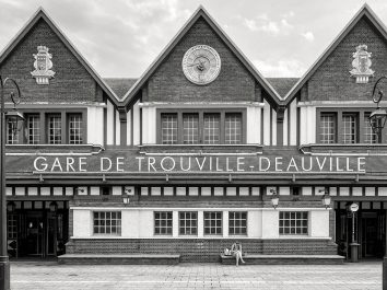Black and white photo of Gare de Trouville-Deauville, a railway station with a clock and symmetrical architecture. A person sits on a bench in front of the building.