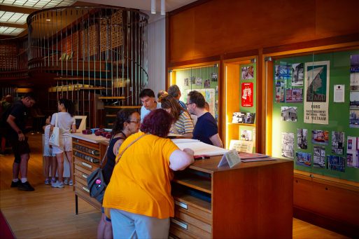 People look at documents and displays in a warmly lit library or archive space, with large wooden cupboards and information panels on the walls, reminiscent of Paris in 1970. A spiral staircase is visible in the background.