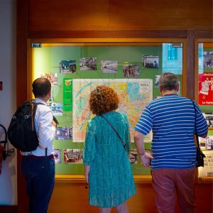 Three people stand in front of a display case studying a large map of Paris, surrounded by postcards and photos from 1970. Their backs are to the camera.
