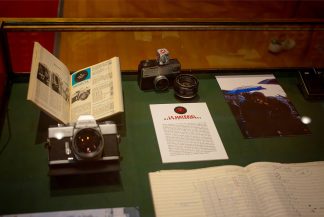 Display case with vintage cameras from 1970, an open book, a typed document, a photograph of a person with an umbrella in Paris, and a handwritten ledger on a green surface. Items are arranged neatly for exhibition.