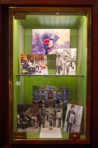 A glass display case with several vintage black-and-white and colour photographs, including candid scenes of Paris in 1970, showing people in various outdoor and street settings, mounted on two shelves against a green background.