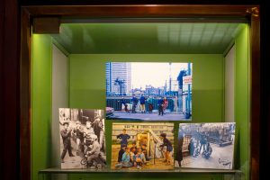 A display case holds four vintage photos of children playing outdoors in urban settings; one colour photo at the back—capturing a lively Paris street scene from 1970—and three black-and-white photos arranged at the front.