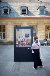 A woman stands in front of an outdoor display featuring a historic photo from Paris, 1970, showing three people walking with luggage, set against an old stone building with windows.