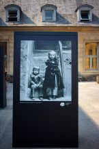 A black-and-white photo of two children, one seated and one standing, is displayed outdoors on a large panel in front of a historic stone building with dormer windows, evoking the timeless streets of Paris in 1970.
