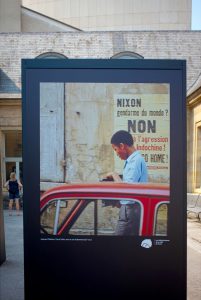 A display board outdoors shows a photo of a man by a red car, beside a French sign protesting Nixon and the Vietnam War. A woman stands in the background, and beige stone buildings surround the scene, capturing the spirit of Paris in 1970.