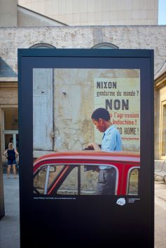 A display board outdoors shows a photo of a man by a red car, beside a French sign protesting Nixon and the Vietnam War. A woman stands in the background, and beige stone buildings surround the scene, capturing the spirit of Paris in 1970.
