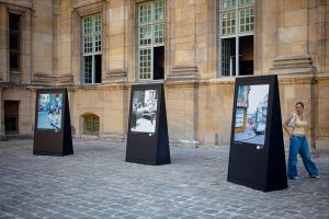 Three large outdoor photo displays stand in front of a historic stone building, evoking the ambience of Paris in 1970. A woman in a yellow top and blue jeans stands nearby, looking at the camera.