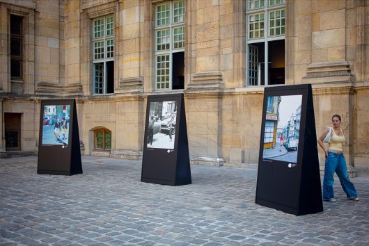 Three large outdoor photo displays stand in front of a historic stone building, evoking the ambience of Paris in 1970. A woman in a yellow top and blue jeans stands nearby, looking at the camera.