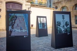 Three large photo displays stand on a cobbled courtyard in Paris. The leftmost image shows children playing; the rightmost, police in riot gear; the middle, a balcony. Echoes of 1970 linger as buildings surround the open area.