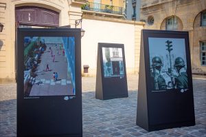 Three large photo displays stand on a cobbled courtyard in Paris. The leftmost image shows children playing; the rightmost, police in riot gear; the middle, a balcony. Echoes of 1970 linger as buildings surround the open area.