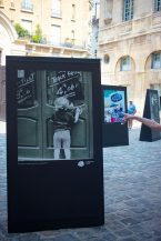 Outdoor art exhibition with black display stands on cobblestones. Foremost display shows a child writing on a door, perhaps evoking the spirit of Paris in 1970. Other artworks are visible behind, along with a person's outstretched arm pointing. Sunlit buildings in background.