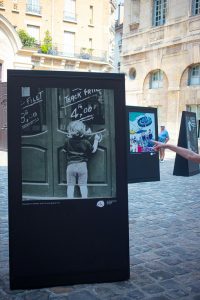 Outdoor art exhibition with black display stands on cobblestones. Foremost display shows a child writing on a door, perhaps evoking the spirit of Paris in 1970. Other artworks are visible behind, along with a person's outstretched arm pointing. Sunlit buildings in background.