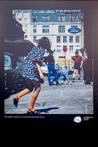 A girl in a patterned dress and orange socks plays hopscotch on a city pavement in Paris, 1970, with parked cars, several people, and a “Ford” parking sign visible in the background.