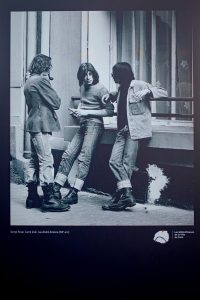 Three young people with rolled-up jeans and boots stand and talk on a city pavement in front of a building, leaning casually. The image is black and white and has a candid, vintage feel reminiscent of 1970s Paris.